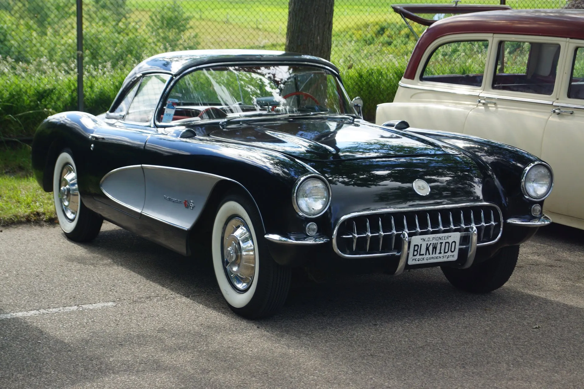 A black 1957 Corvette with silver side coves and its black hardtop fitted, parked on asphalt and seen from the front corner, with Fuel Injection script and crossed flags on the cove.