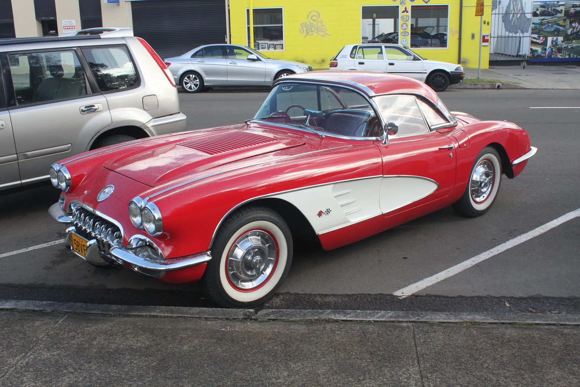 A red 1958 Corvette with white side coves and a red hardtop parked at a curb, seen from the front corner, showing the ribbed louver panel on its hood and four headlights.