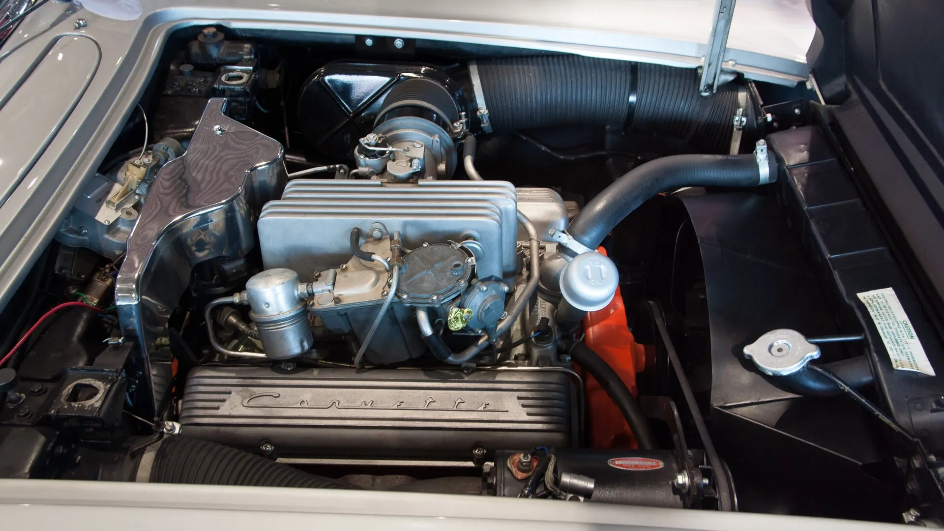 The engine bay of a silver C1 Corvette with a fuel-injected V8: a finned aluminum fuel injection unit, a large black intake duct and a finned valve cover with Corvette script.