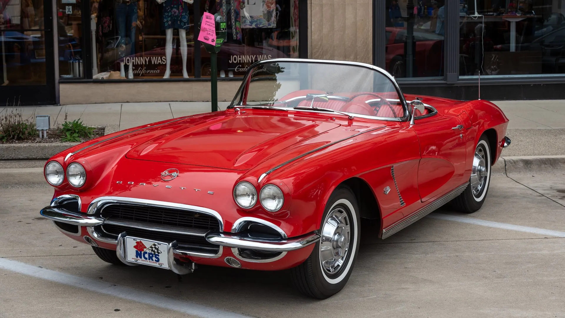 A red 1962 Corvette roadster with its top down, parked at a curb in front of a shop window, with one-color side coves, ribbed rocker trim and four headlights.
