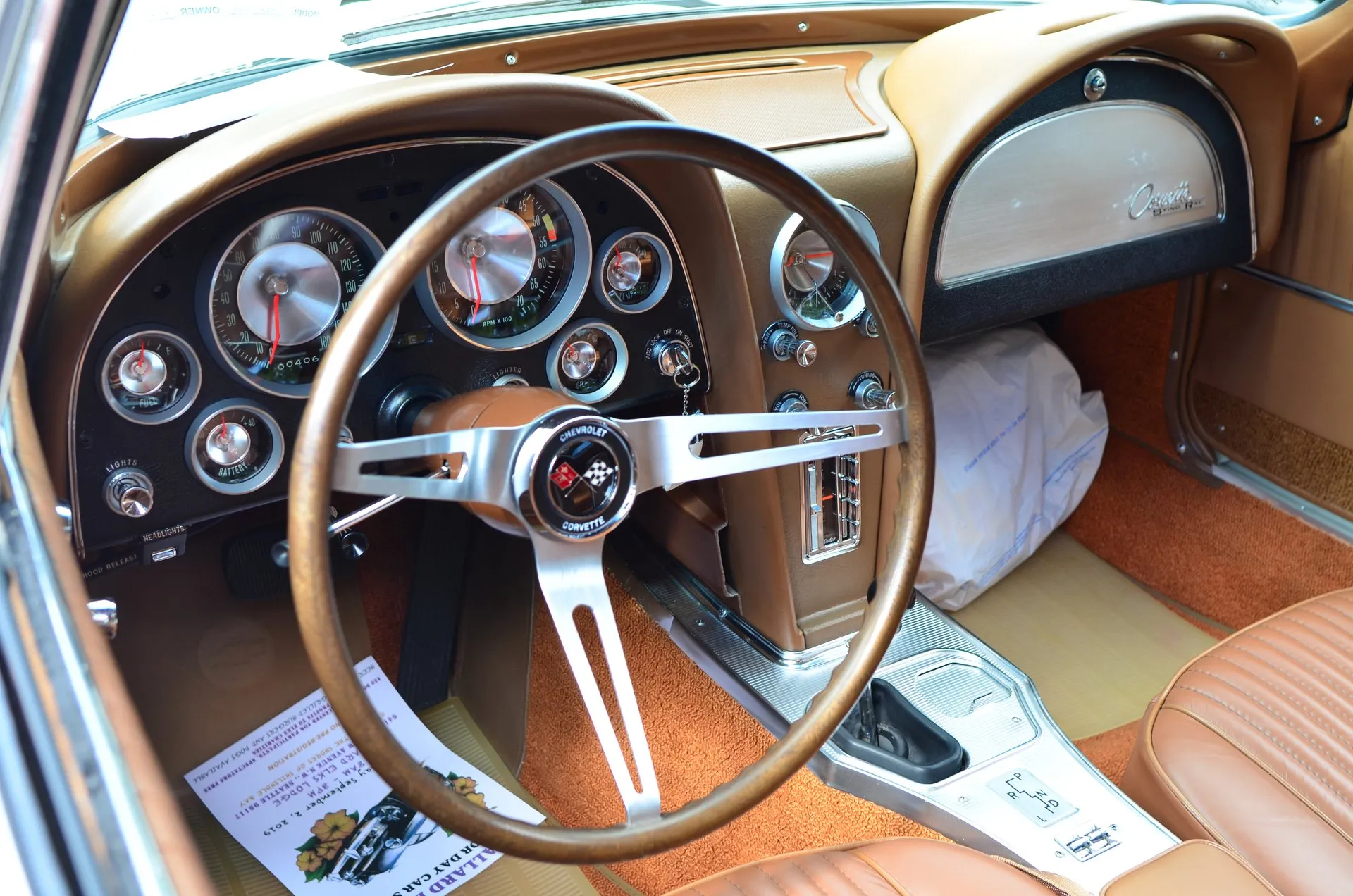 The dashboard of a Corvette coupe with a saddle interior, seen through the open driver's door: two hooded cowls, a speedometer and tachometer behind a wood-rim steering wheel, and an automatic shift pattern on the center console.