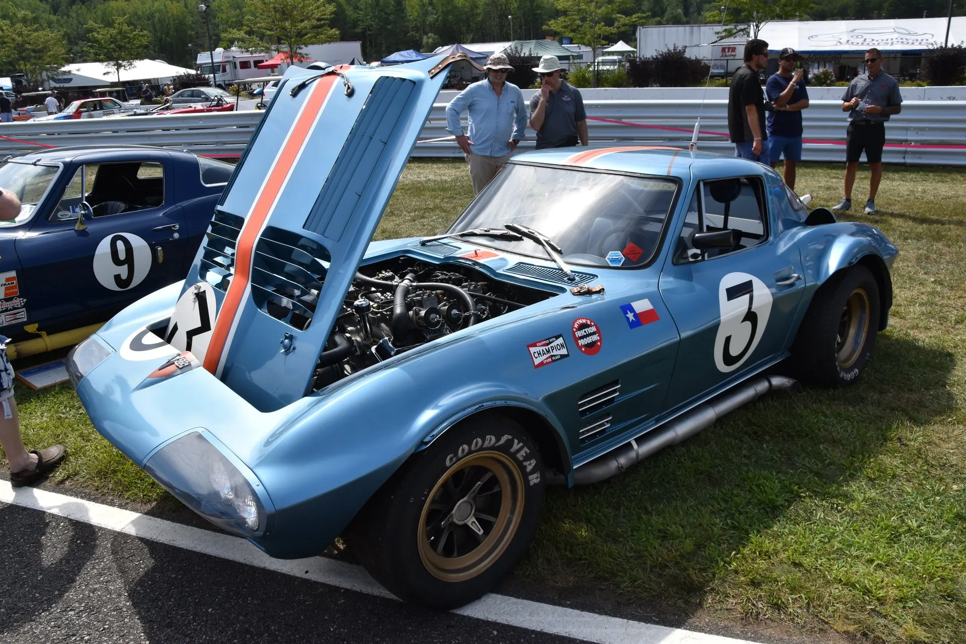 A light blue 1963 Corvette Grand Sport race coupe with an orange stripe and the number 3, its front-hinged hood raised over the V8 and side exhaust pipes under the door, parked on grass beside a racetrack with spectators behind it.