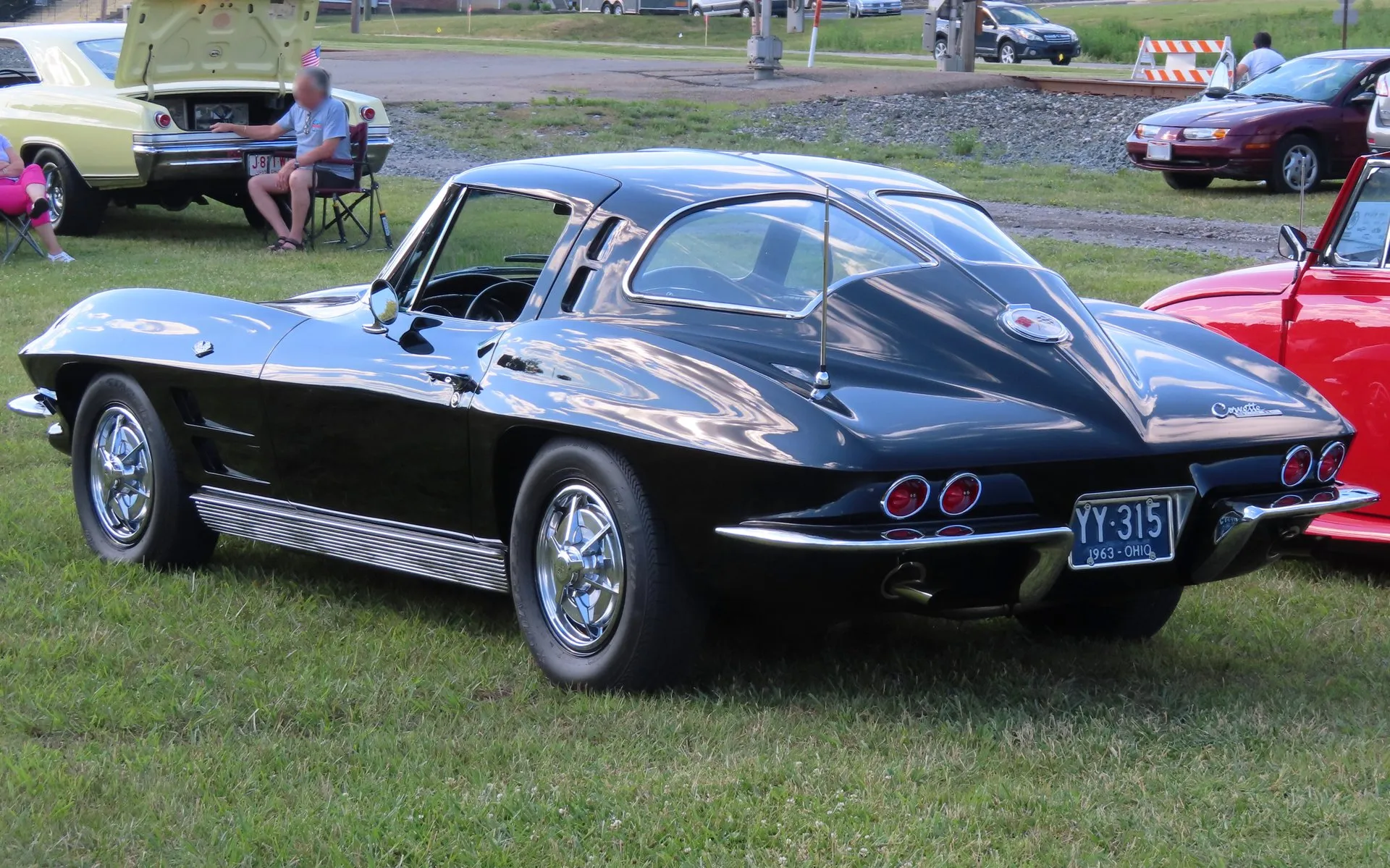 A black 1963 Corvette Sting Ray coupe parked on grass at a car show, seen from the rear left, with a body-color bar splitting the rear window down the middle, ribbed chrome rocker trim and a 1963 Ohio license plate.