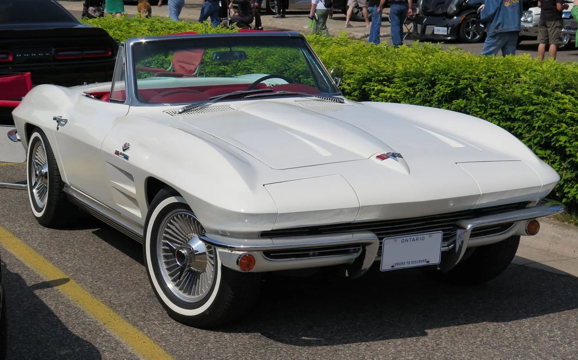 A white 1964 Corvette convertible with its top down and a red interior, parked at a car show and seen from the front right, with whitewall tires and two long recesses in the hood.