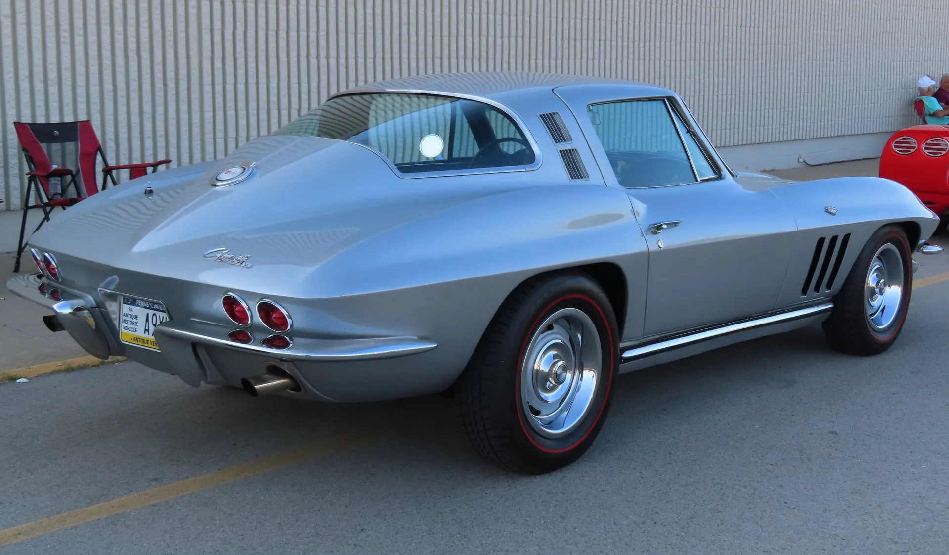 A silver 1965 Corvette coupe in a parking lot, seen from the rear right, with a one-piece rear window, vents in the roof pillar behind the door, three vertical vents in the front fender, and Rally wheels with red-stripe tires.