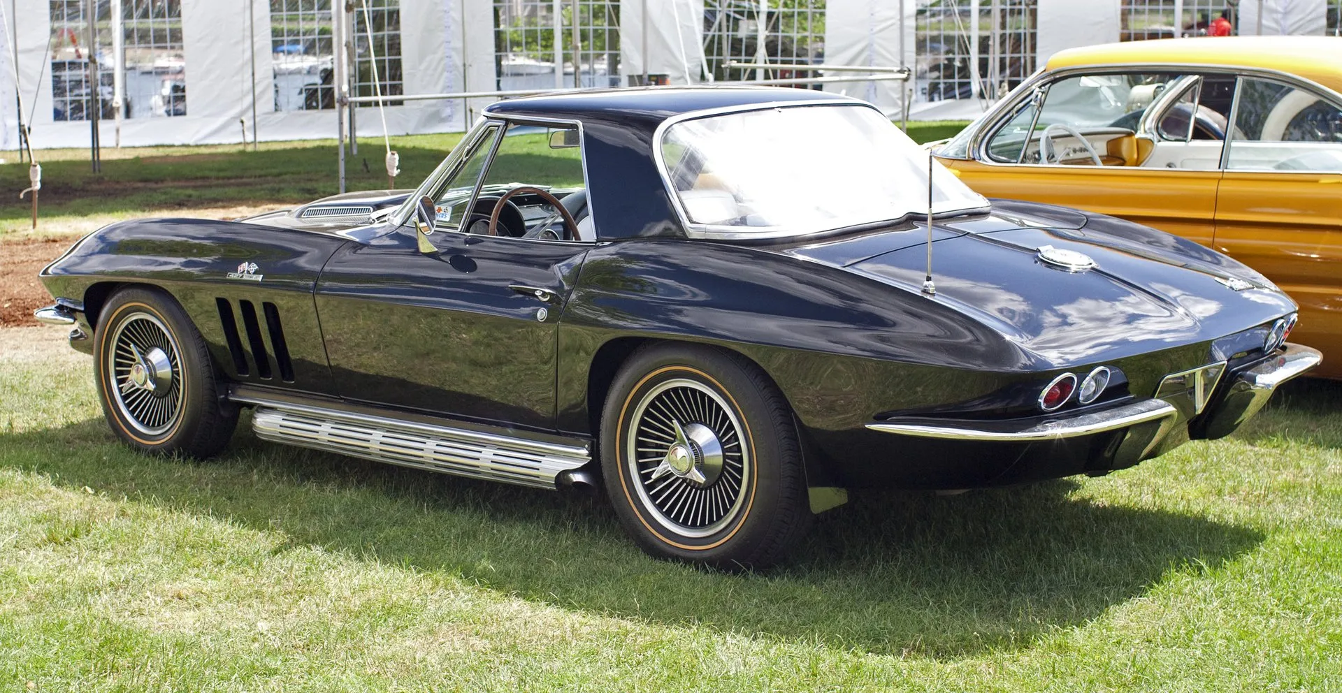 A black 1966 Corvette convertible wearing its removable hardtop, parked on grass and seen from the rear left, with chrome side exhaust covers below the doors, gold-stripe tires, three fender vents and a 427 badge under the crossed-flags emblem.