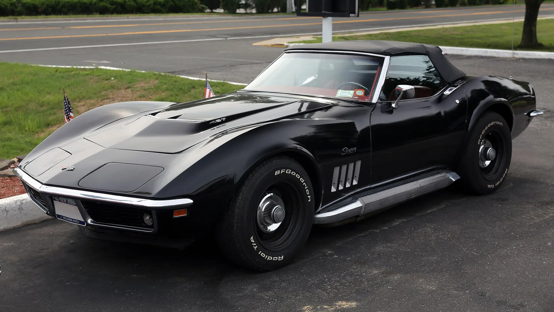 A black 1969 Corvette Stingray convertible with its soft top up and side exhaust pipes, parked on asphalt and seen from the front left, with a 427 badge on the hood.
