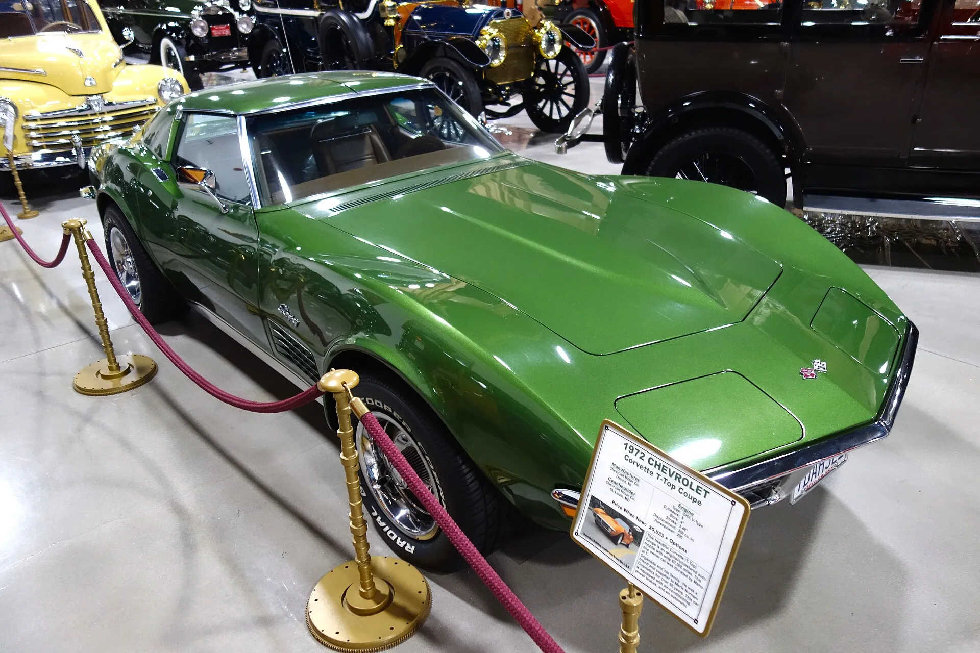 A green 1972 Corvette coupe on display in a museum behind rope barriers, seen from above the front right, with an information placard in front of it.