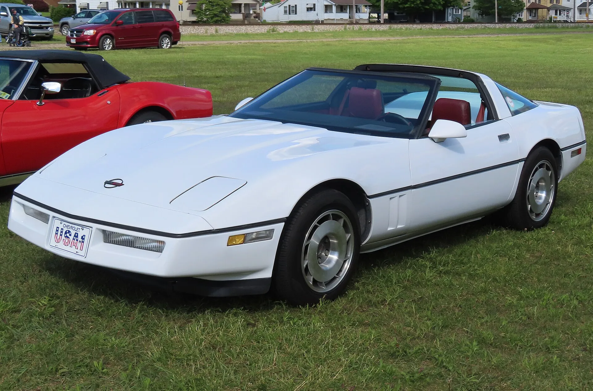 White 1984 Chevrolet Corvette coupe with its roof panel off and a red interior, parked on grass at a car cruise, seen from the front left.