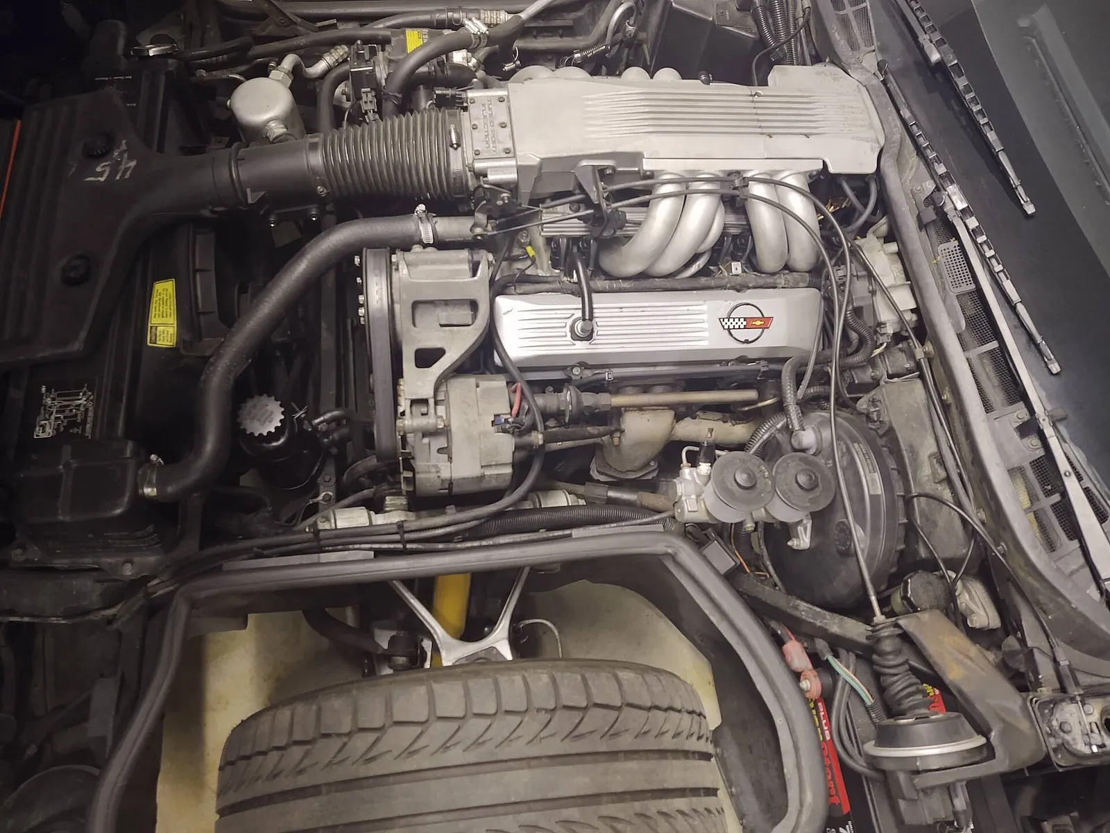 Engine bay of a 1985 C4 Corvette seen from the driver side, with the front tire at the bottom, the brake booster at right and a side-terminal battery tucked between the wheel well and the firewall.