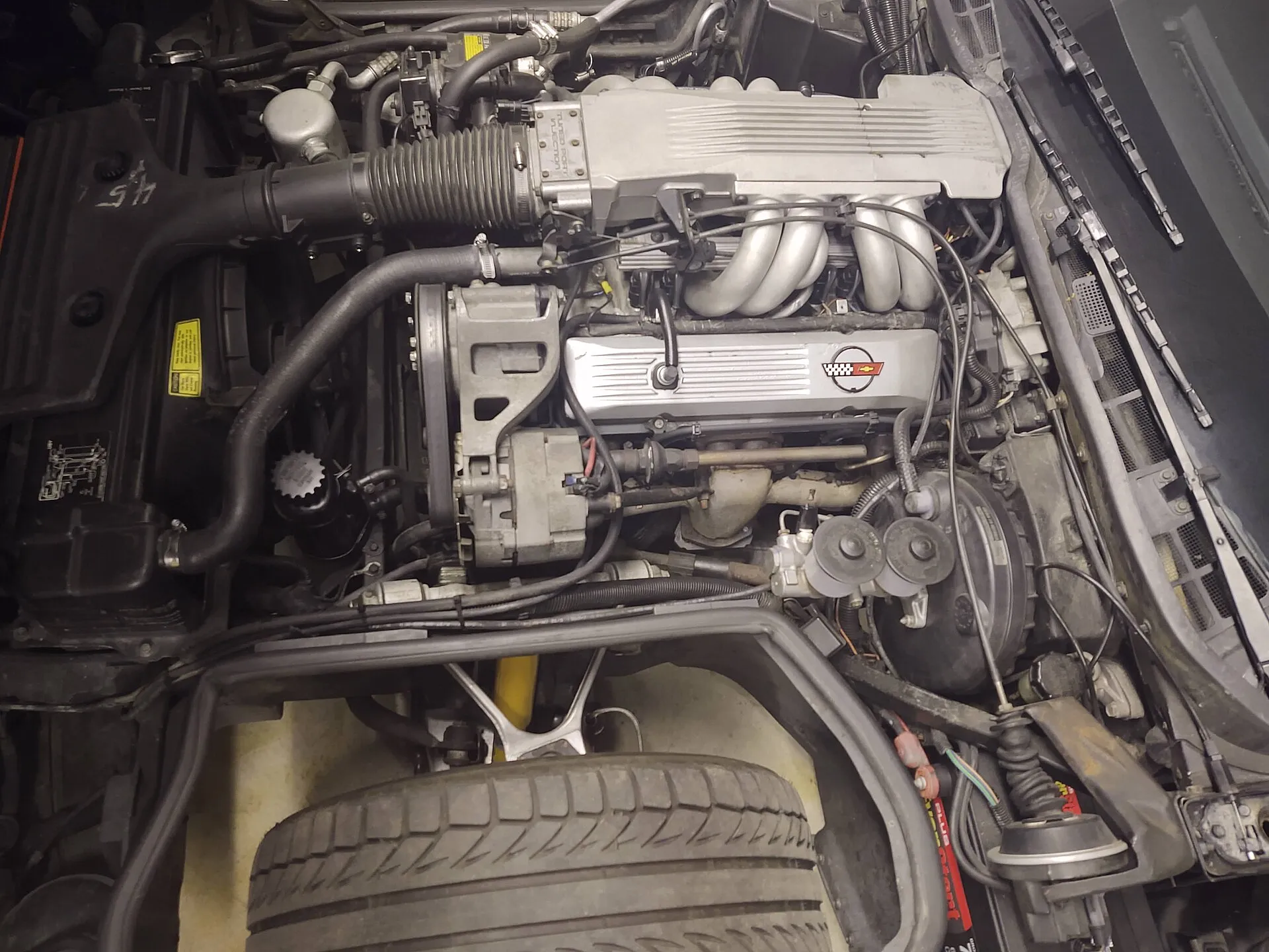 Engine bay of a 1985 Corvette seen from the driver side, showing the silver tuned-port intake plenum and runners on top of the V8, with the front tire at the bottom of the frame.