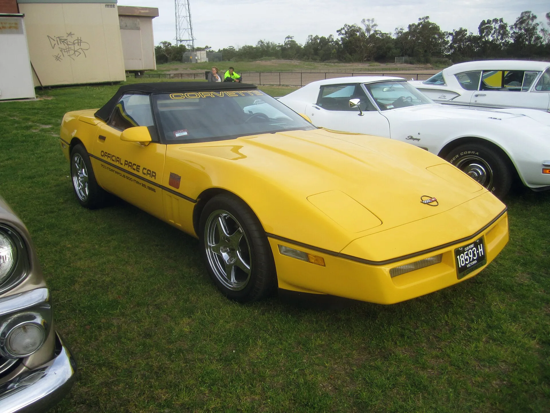 Yellow 1986 Chevrolet Corvette convertible with its black top up and Official Pace Car lettering on the door, parked on grass at a car show.