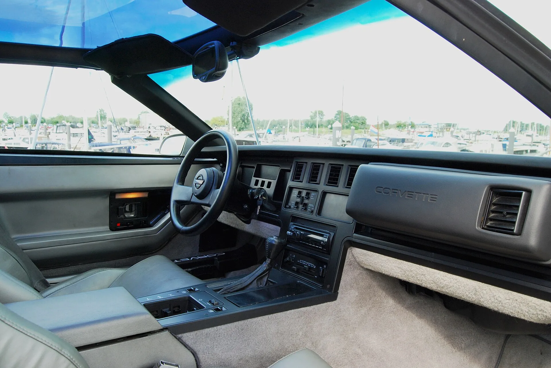 Interior of a 1986 Corvette coupe seen from the passenger side, with gray leather seats, the steering wheel and the angular dashboard and center console.