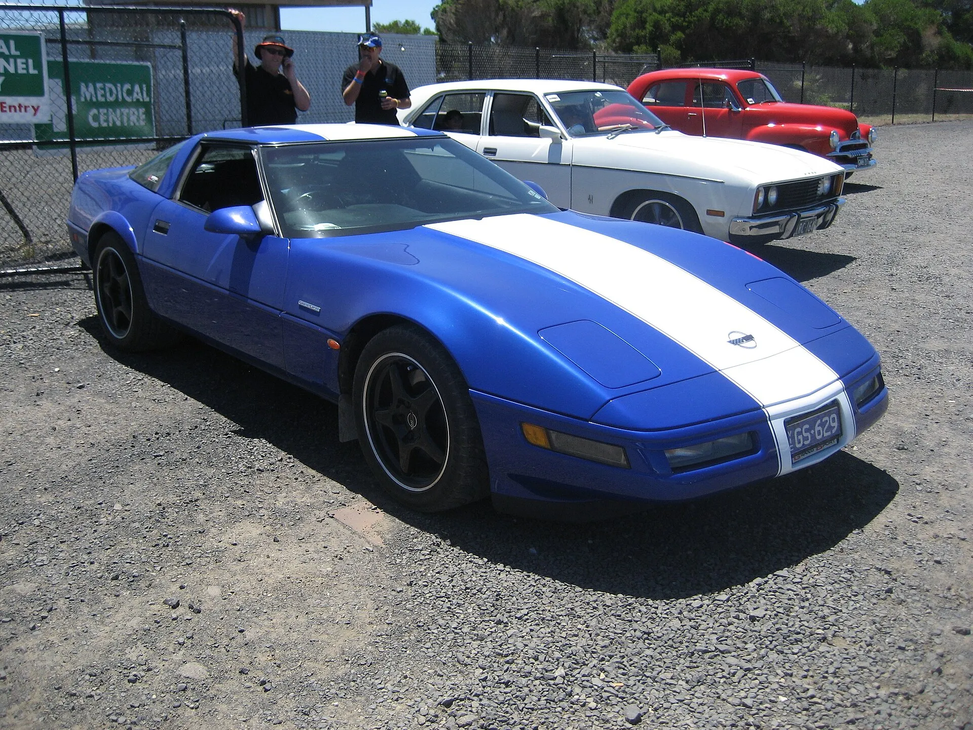Blue 1996 Chevrolet Corvette Grand Sport coupe with a white center stripe and black wheels, parked on gravel at a car show.