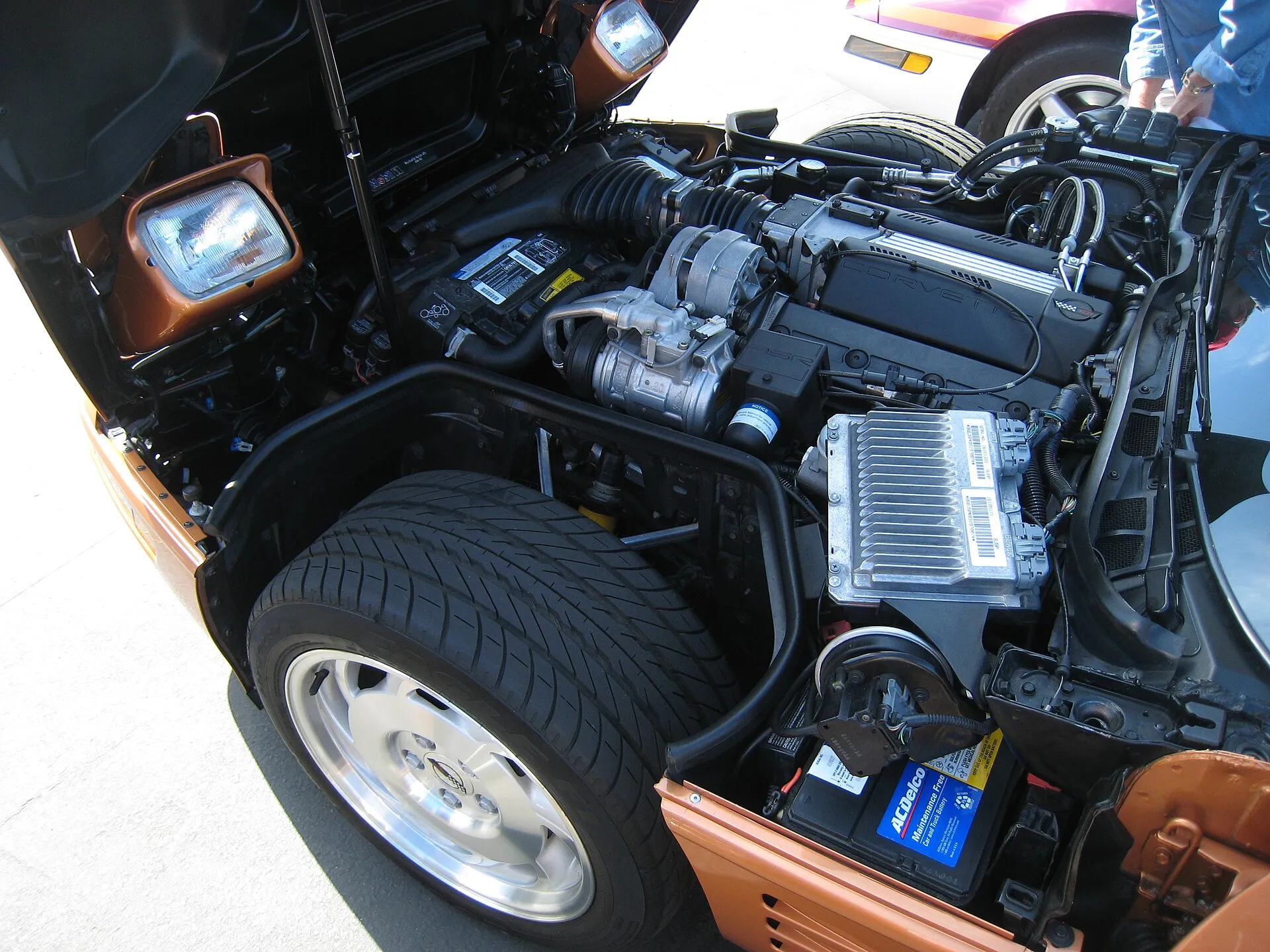 Engine bay of a copper C4 Corvette seen from the driver side with the clamshell hood up; the battery sits at the bottom right, just behind the front tire.
