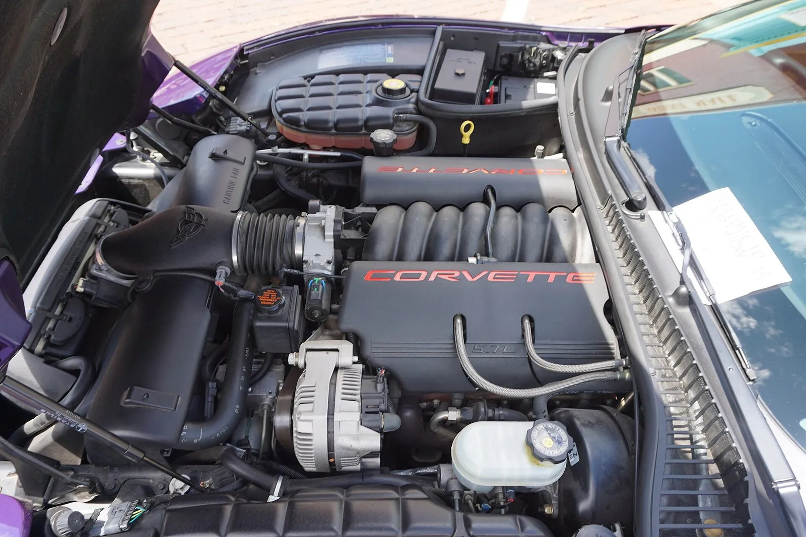 Engine bay of a purple 1998 C5 Corvette with the hood up; at the far corner by the windshield, a black fuse box sits beside the battery and its red terminal cover.