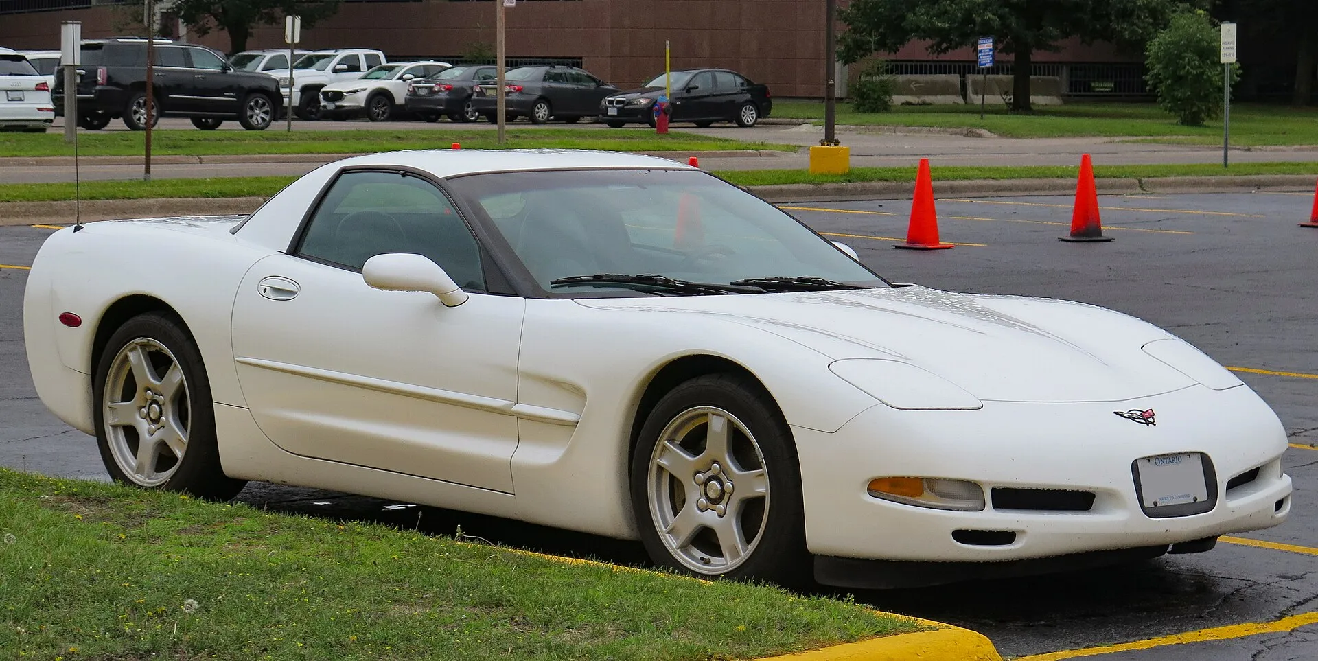 White 1999 Chevrolet Corvette C5 hardtop, the fixed-roof model, parked in a lot and seen from the front right, with orange traffic cones behind it.