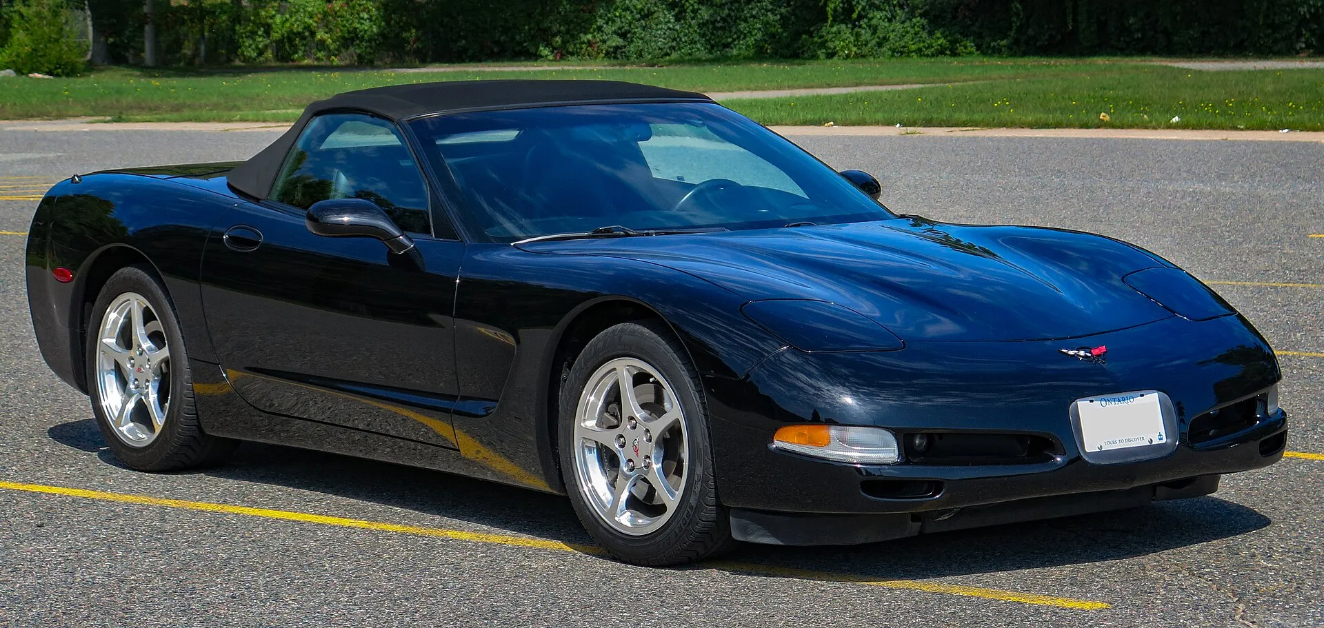 Black 2002 Chevrolet Corvette C5 convertible with its black soft top up, parked in an empty lot and seen from the front right, with polished five-spoke wheels.