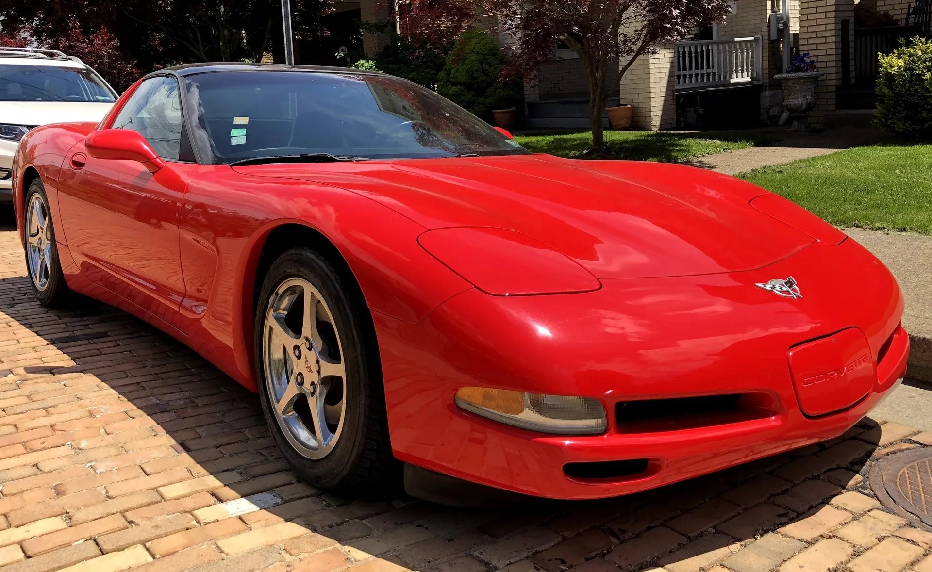 Red 2003 Chevrolet Corvette C5 coupe with its headlights folded down, parked on brick paving beside a lawn and seen from the front right, with silver five-spoke wheels.