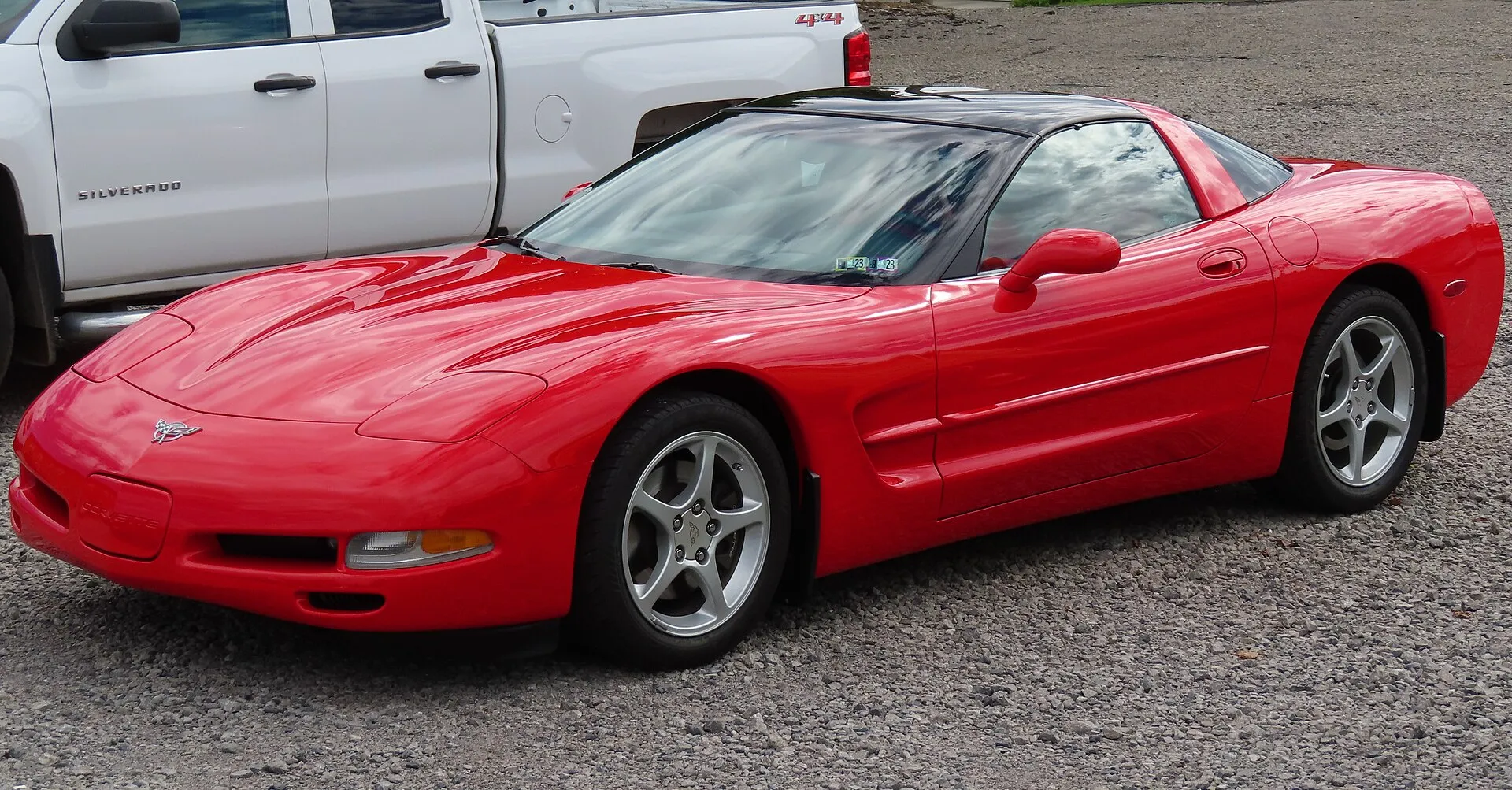 Red 2003 Chevrolet Corvette C5 coupe with a dark roof panel, parked on gravel and seen from the front left, with a white pickup truck parked behind it.