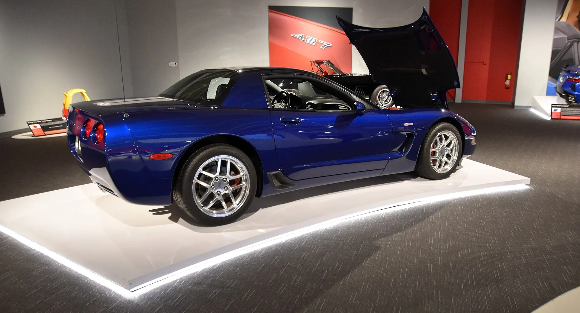 Blue 2004 Corvette Z06 Commemorative Edition on a museum display platform, seen from the rear left with the hood raised and a silver stripe on the rear deck.