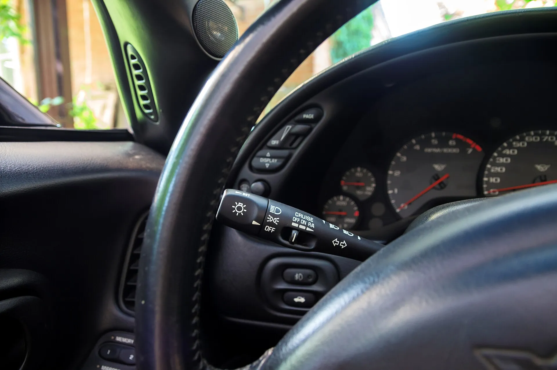 Driver's view of a C5 Corvette cockpit: leather steering wheel, the turn signal stalk with headlight and cruise control settings, a switch panel left of the gauges marked PAGE, OFF and DISPLAY, and analog gauges with red needles.