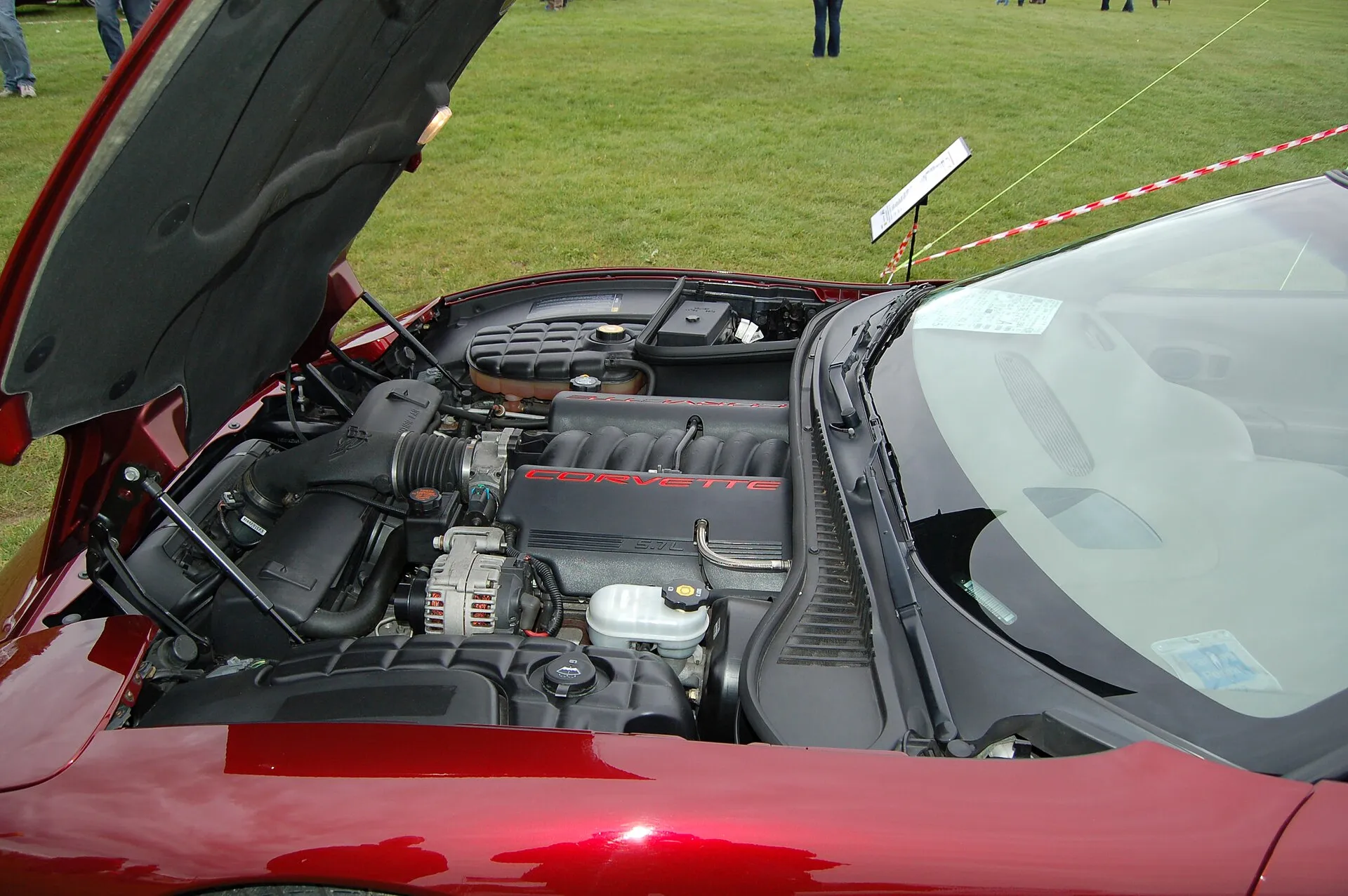 Engine bay of a dark red C5 Corvette with the hood open, showing the black LS1 V8 intake with red Corvette lettering, the air intake duct and a white fluid reservoir.