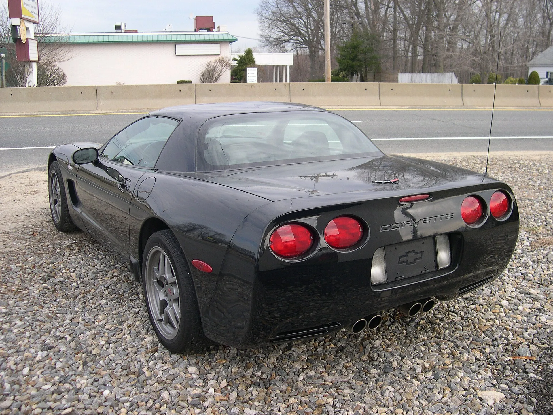 Black 2002 Chevrolet Corvette Z06 on a gravel lot, seen from the rear left, showing the fixed roof with a trunk lid instead of a glass hatch, four round taillights, a fixed radio antenna, gray five-spoke wheels and four exhaust tips.
