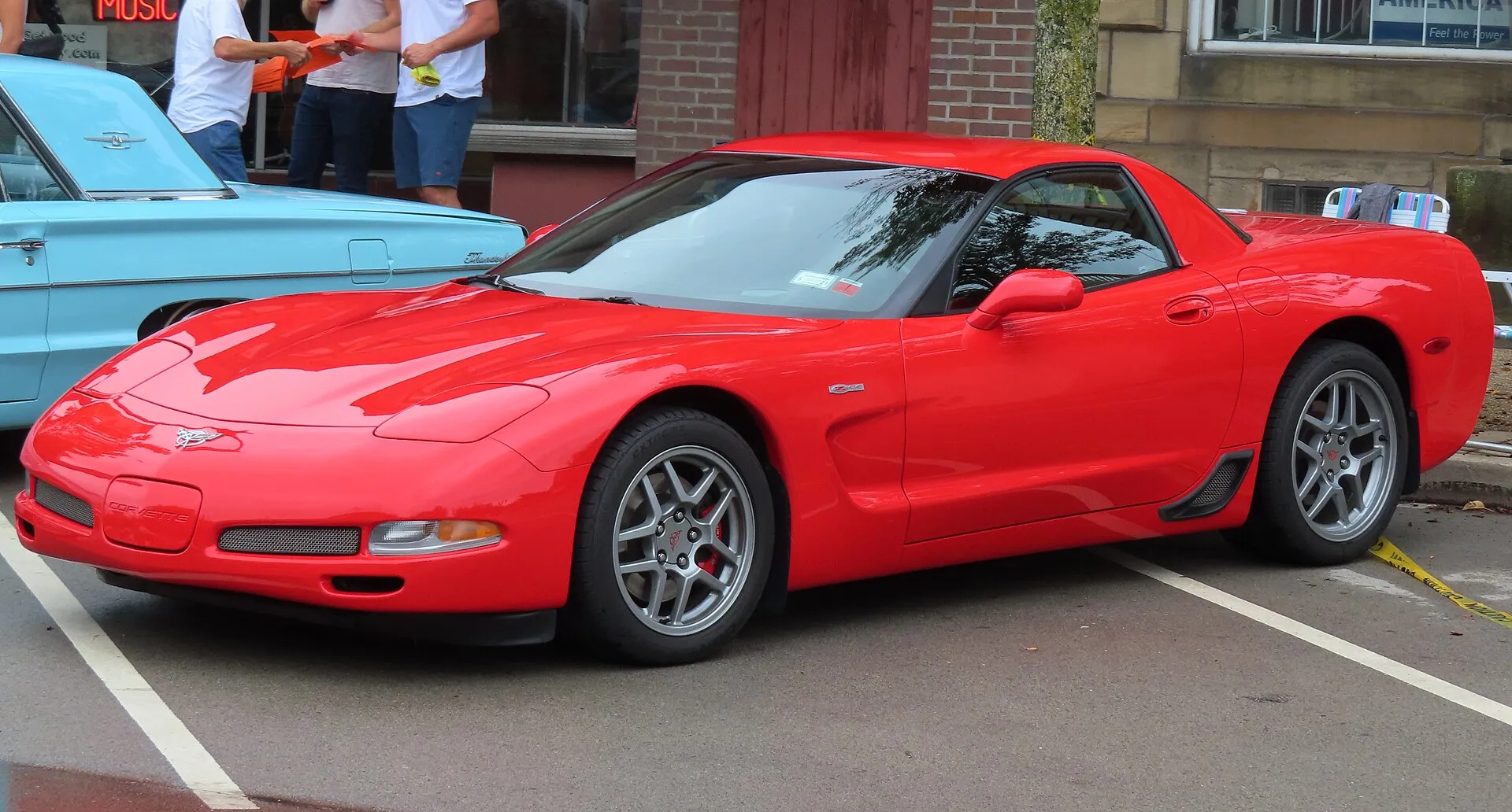 Red 2003 Chevrolet Corvette Z06 parked at a street car show, seen from the front left, with silver split-spoke wheels, red brake calipers and a Z06 badge on the front fender.
