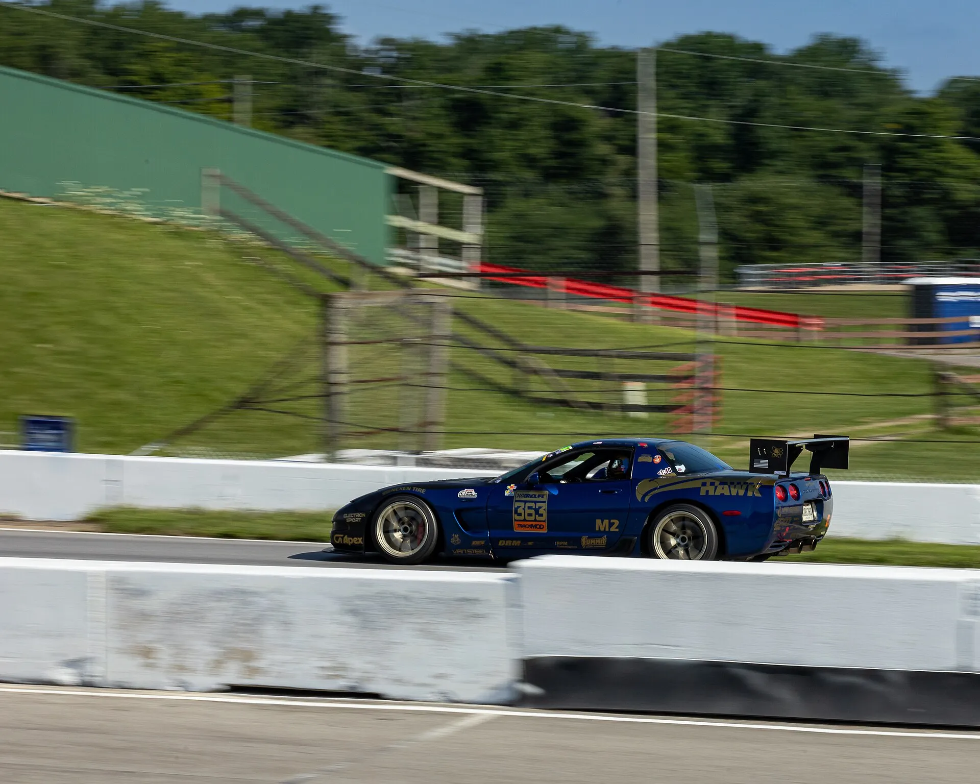 Blue 2003 Chevrolet Corvette Z06 with a large rear wing and sponsor decals, car number 363, driving past concrete barriers at Mid-Ohio during an SCCA time trial.