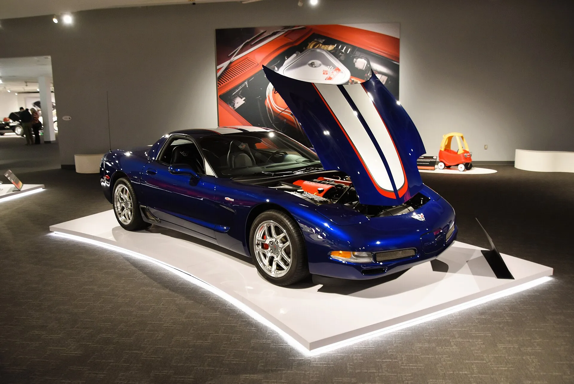 Le Mans Blue 2004 Corvette Z06 Commemorative Edition on a lit museum display platform, seen from the front right with the hood raised to show red engine covers, and a silver and red stripe running over the hood and roof.