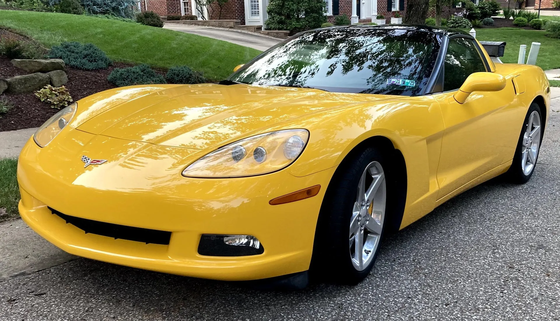 Yellow 2005 Chevrolet Corvette C6 coupe with a black roof panel, parked on a residential street and seen from the front left, with exposed headlights and silver five-spoke wheels.