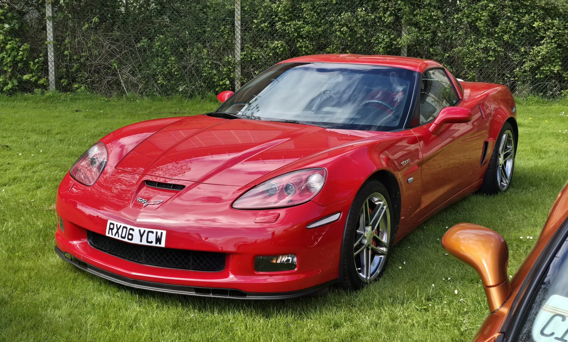 Red 2006 Chevrolet Corvette Z06 parked on grass in front of a hedge, seen from the front left, with silver split-spoke wheels, red brake calipers, a front splitter and a Z06 badge on the front fender.