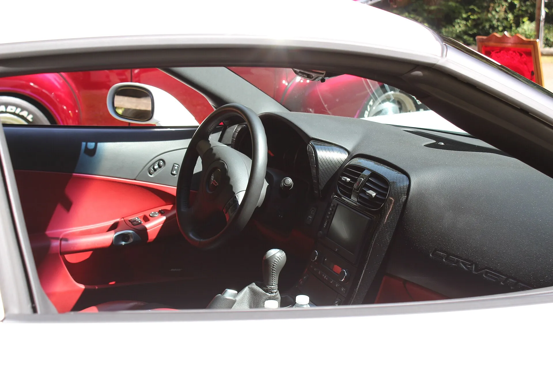 View through the passenger-side window of a 2007 Chevrolet Corvette Z06 cabin, with red door panels, a black dash, the steering wheel and a six-speed manual shifter