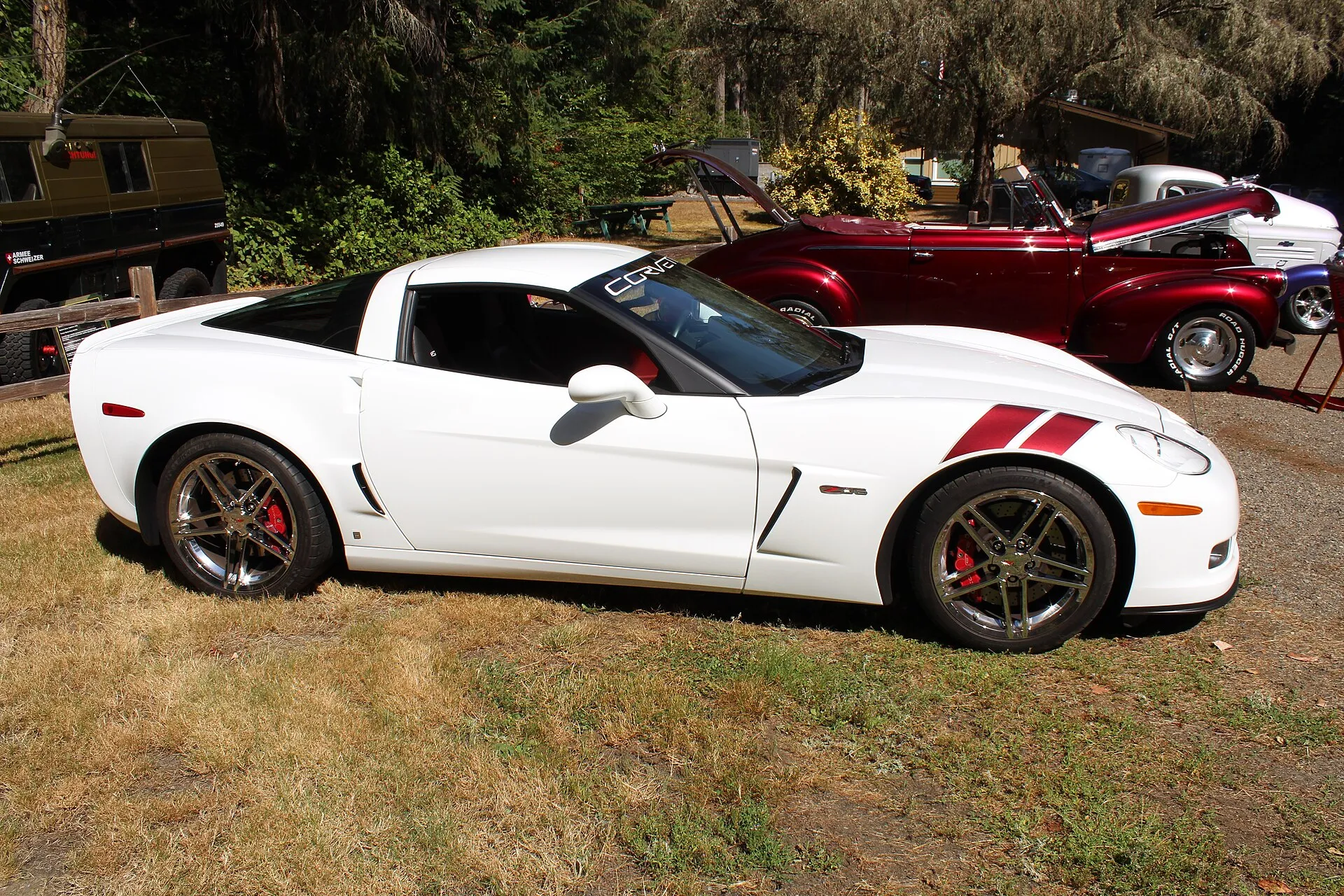 White 2007 Chevrolet Corvette Z06 in side profile with dark red fender stripes and chrome wheels, parked on grass at a car show
