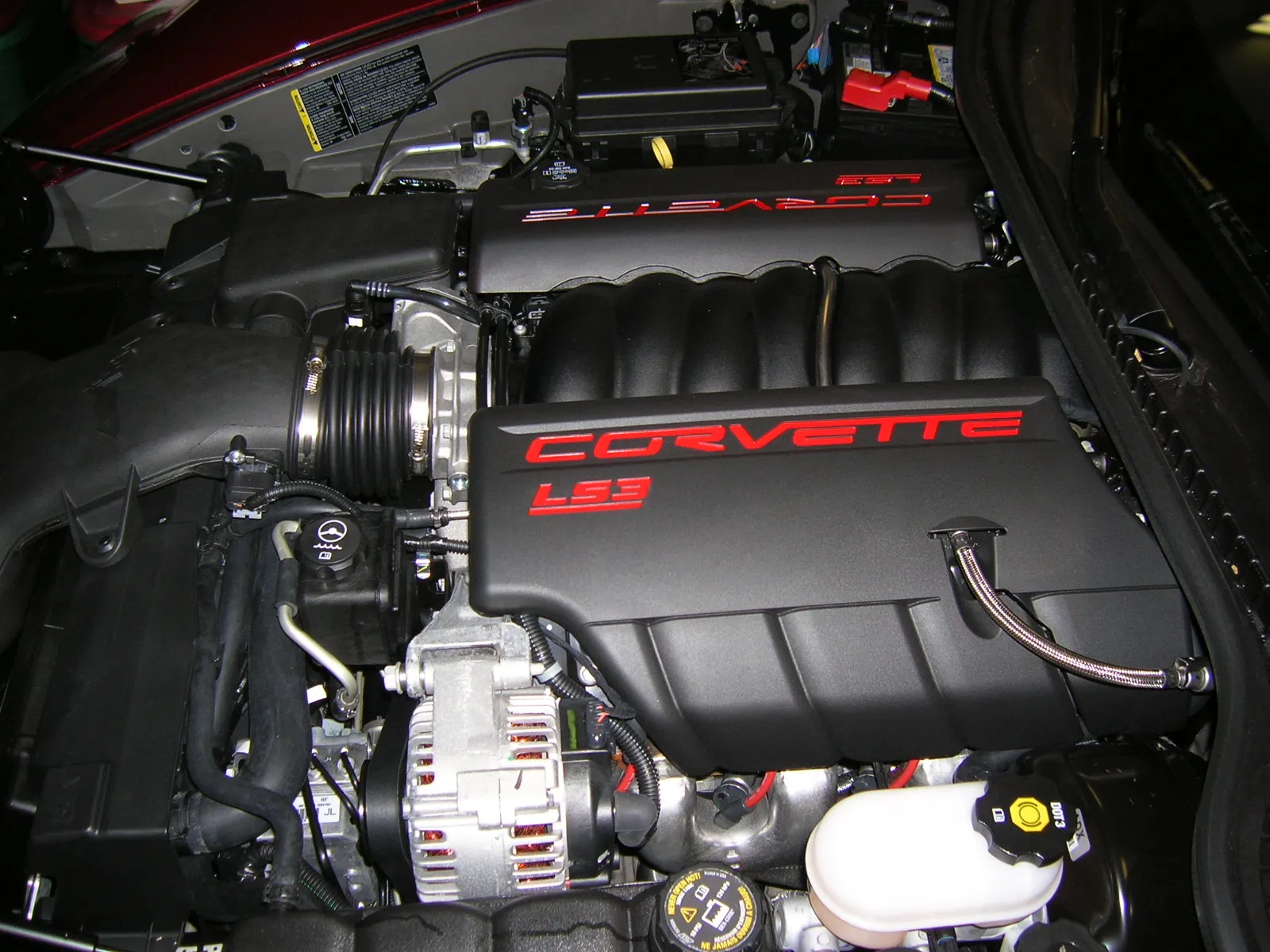 Engine bay of a 2008 C6 Corvette with a black LS3 engine cover; at the top, against the cowl, the black underhood fuse block sits next to the battery with a red positive terminal cover.