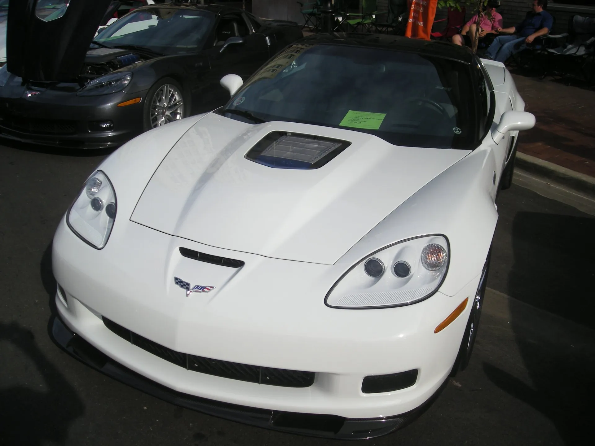 White 2010 Chevrolet Corvette ZR1 with a black carbon fiber roof and a clear window in the hood, parked at an outdoor car show, with a dark gray Corvette behind it, its hood raised.