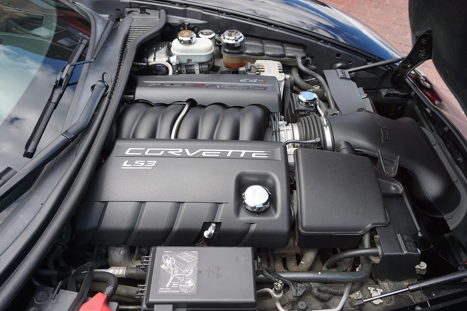 Engine bay of a 2011 C6 Corvette with an LS3 engine cover; in the foreground, the underhood fuse block cover carries a printed jump-start diagram.
