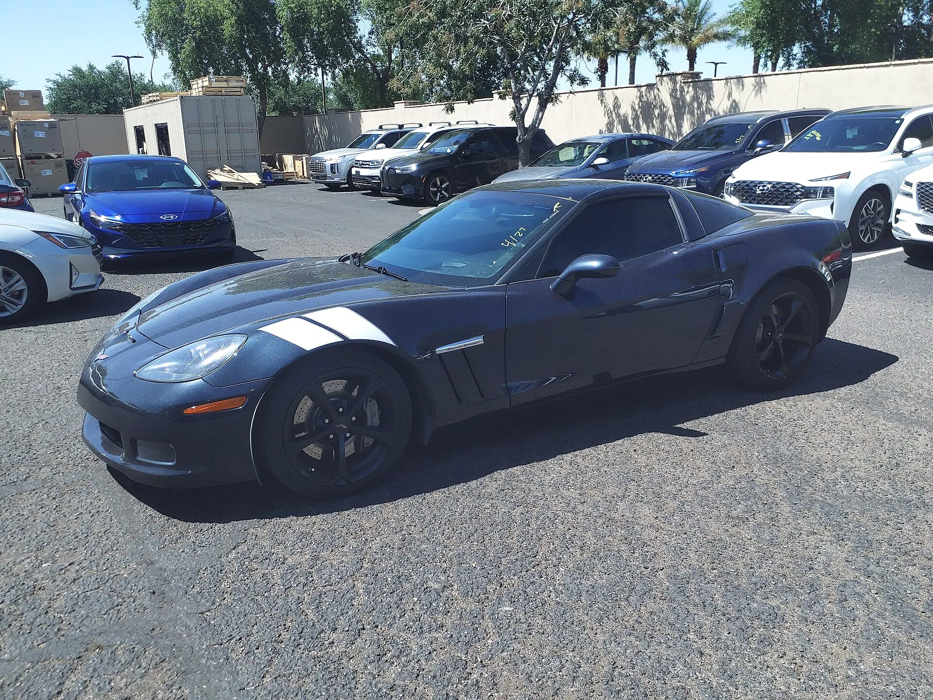 Dark blue 2013 Chevrolet Corvette Grand Sport coupe with white fender stripes on a dealer lot, with grease-pencil marks on the windshield