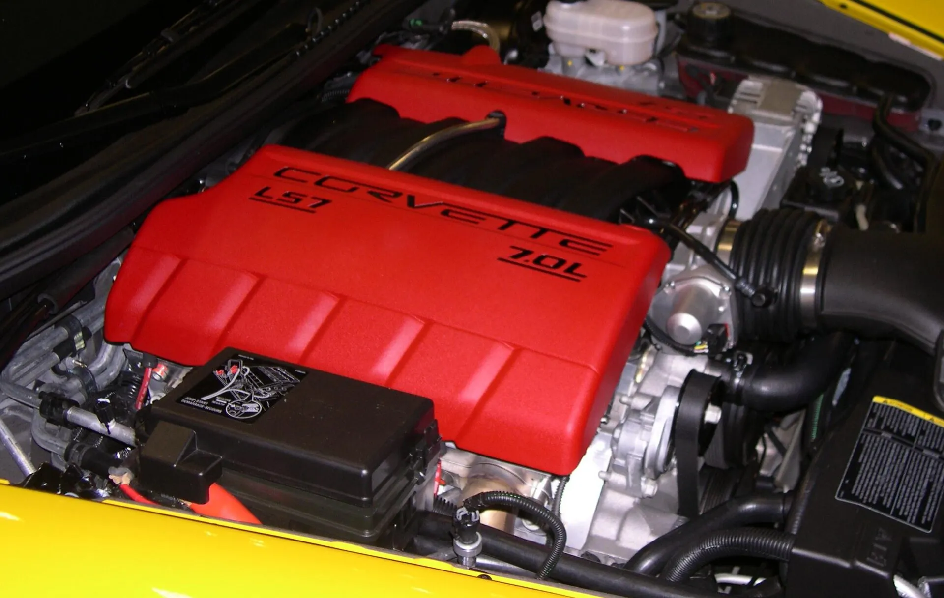 Engine bay of a yellow 2006 C6 Corvette Z06 with a red LS7 engine cover; in the foreground, the black underhood fuse block carries a printed jump-start diagram and a thick red cable.