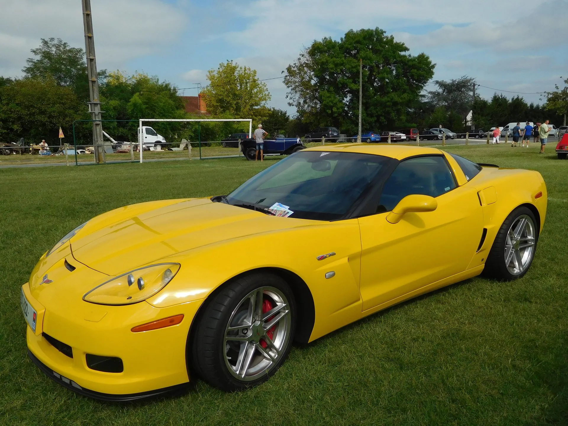 Yellow Chevrolet Corvette Z06 coupe on the grass at an outdoor car show, seen from the front left, with silver split-spoke wheels, red brake calipers, a Z06 badge on the front fender and a flyer at the base of the windshield.