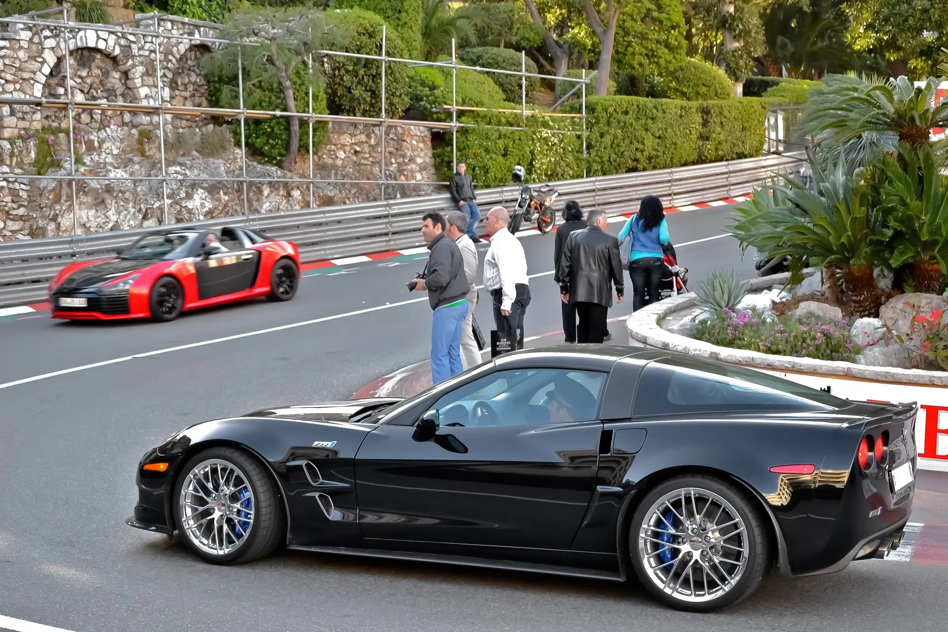 Black Chevrolet Corvette ZR1 seen from its left side on a street, with a ZR1 badge and two vents on the front fender, chrome multi-spoke wheels and blue brake calipers, and people standing at the roadside behind it.