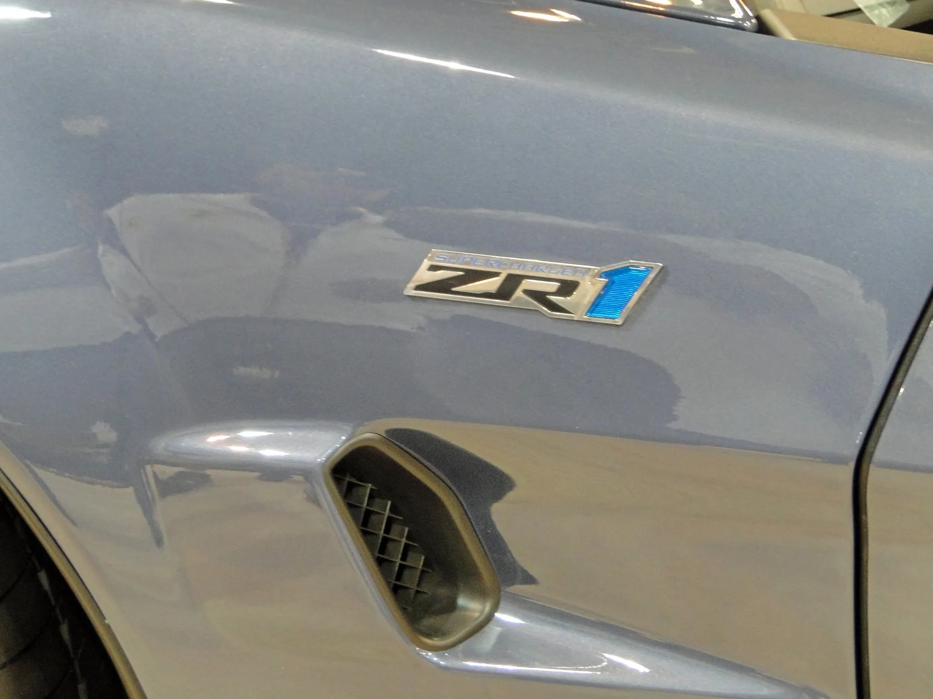 Close-up of the front fender of a blue-gray Corvette ZR1, with a ZR1 badge marked SUPERCHARGED above a vent in the fender.