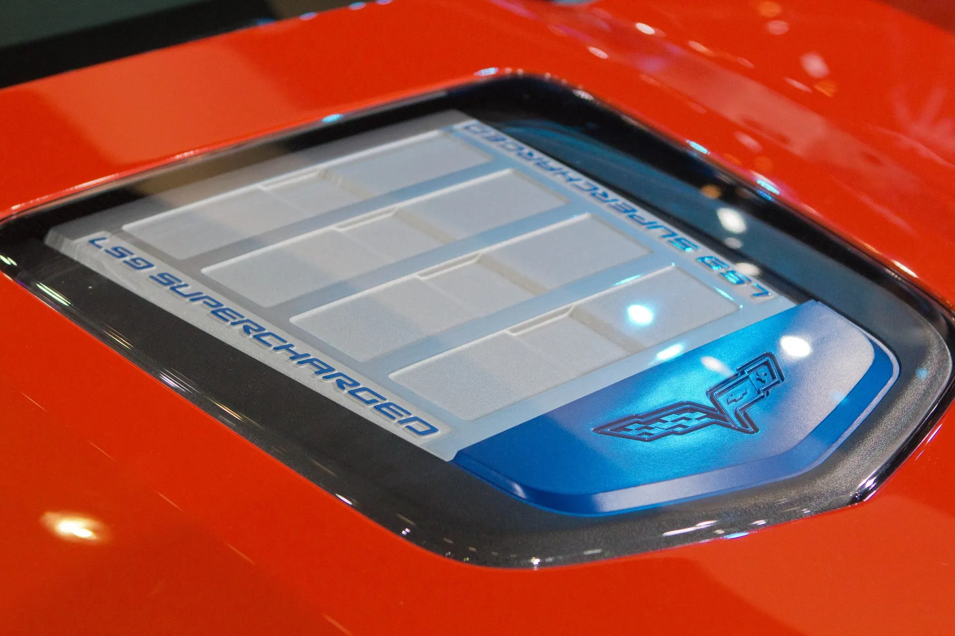 Close-up of the clear window in a red Corvette ZR1's hood, showing a silver cover lettered LS9 SUPERCHARGED in blue and a blue panel with the crossed-flags emblem.