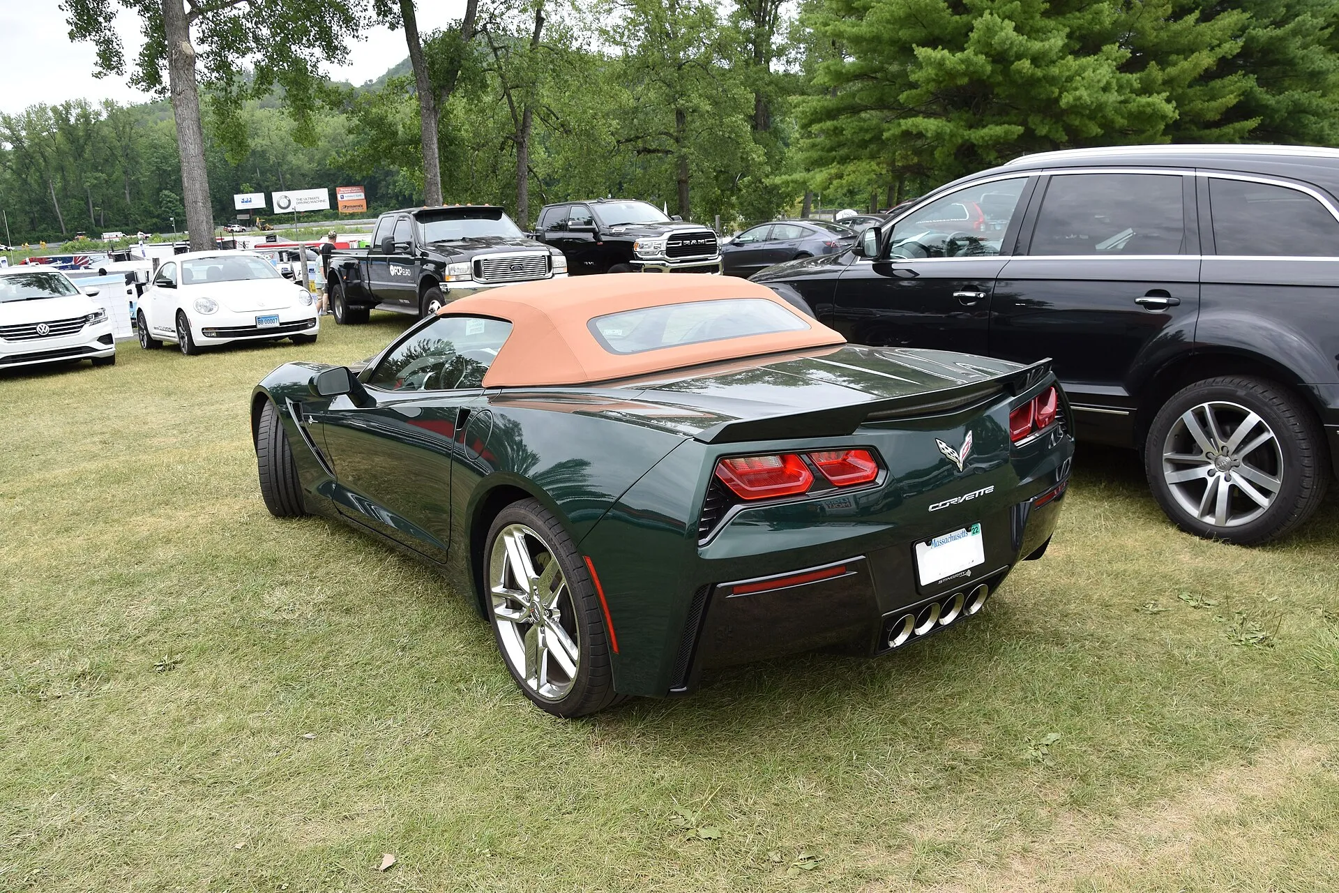 Dark green 2014 Chevrolet Corvette Stingray convertible with a tan soft top and chrome wheels, seen from the rear left on grass at a car show.