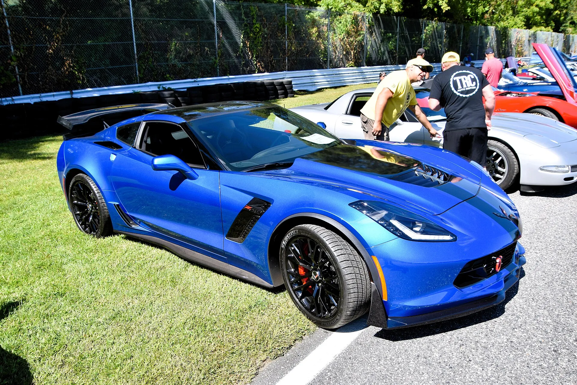 Blue C7 Chevrolet Corvette Z06 coupe with black wheels, red brake calipers and a rear wing, seen from the front right at a car show beside a racetrack.