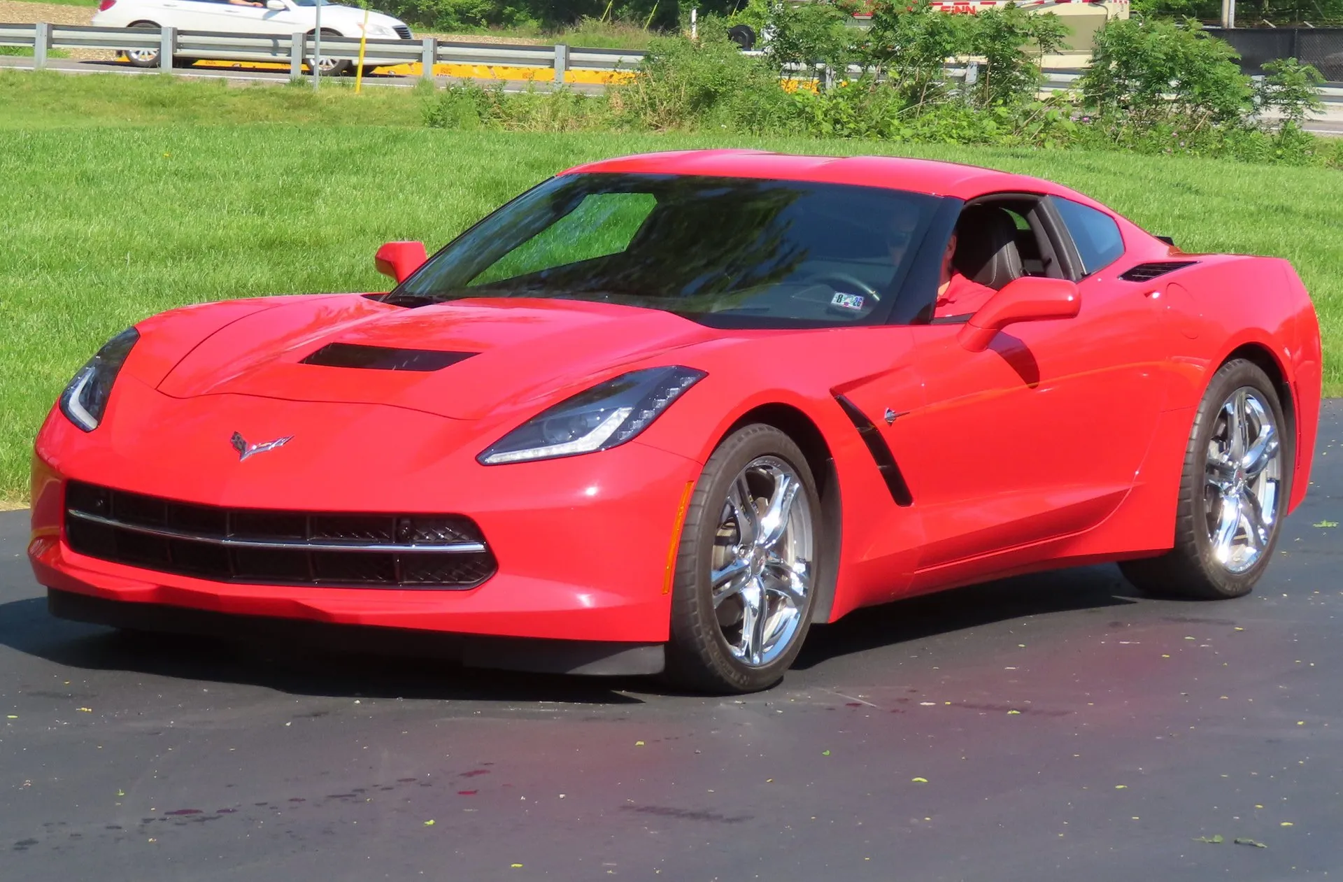 A red 2016 C7 Corvette Stingray coupe with chrome wheels on a paved lot beside grass and a guardrail, seen from the front left.