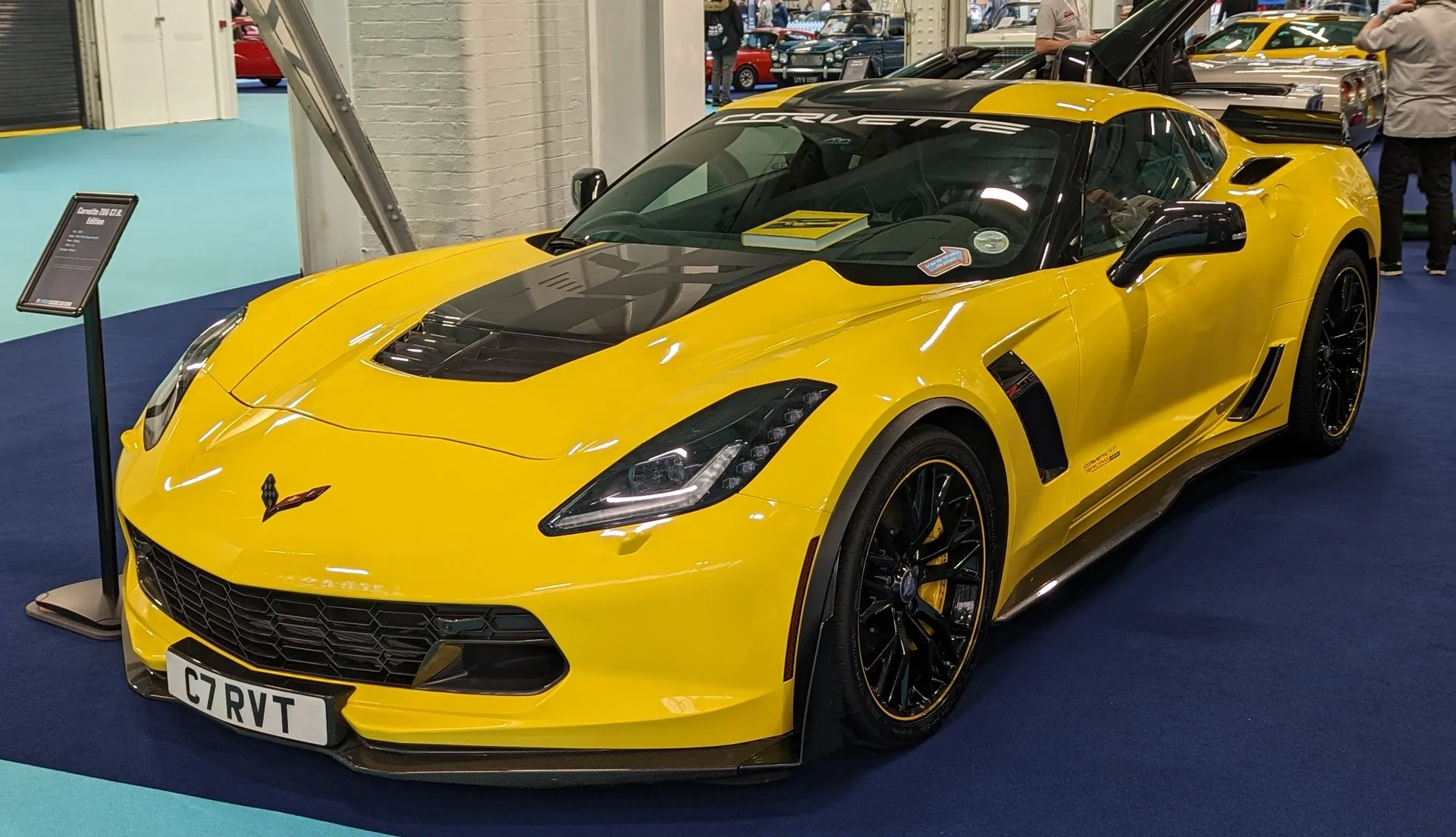 Yellow 2016 Corvette Z06 C7.R Edition coupe with a black hood center and black wheels with yellow rim stripes, on display at an indoor show beside an information stand, seen from the front left.