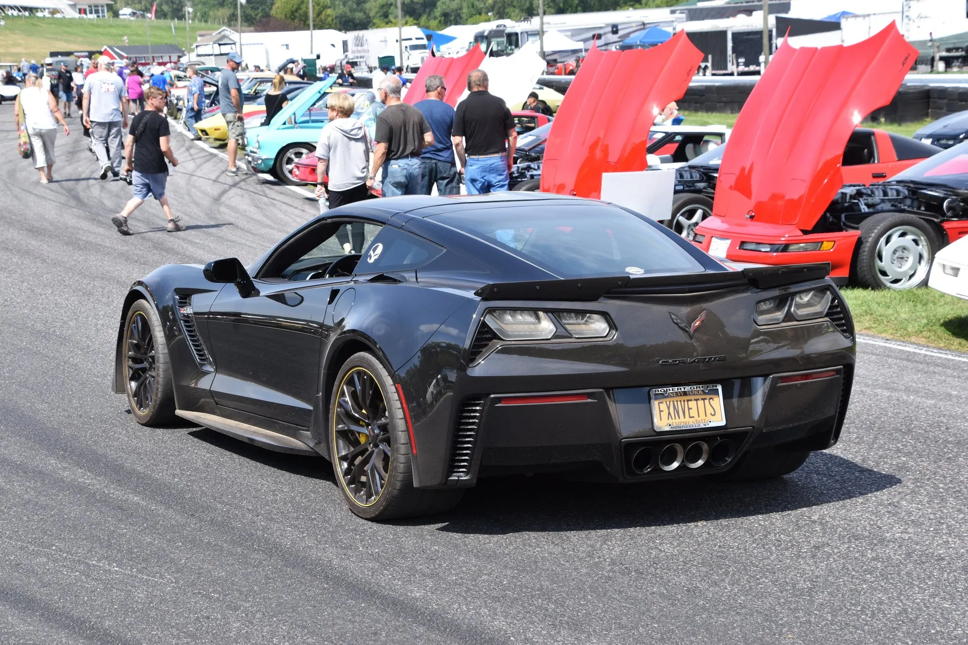 Dark gray C7 Corvette Z06 coupe on the asphalt at a car show at Lime Rock Park, seen from the rear left, with four center exhaust tips, a rear spoiler, wide rear fenders and black wheels with a gold rim stripe, spectators and other Corvettes behind it.
