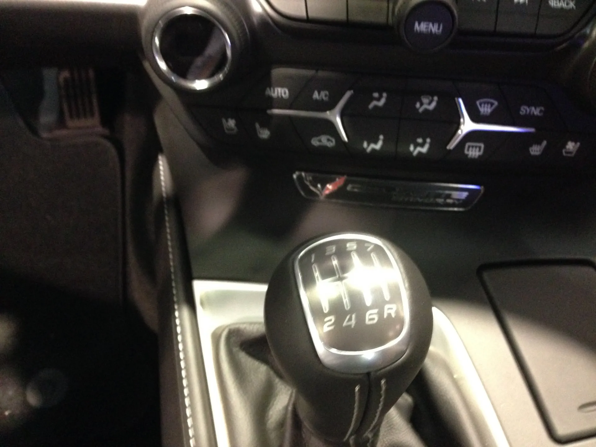 The shift knob of a C7 Corvette Stingray's seven-speed manual, its pattern reading 1 3 5 7 over 2 4 6 R, below the climate controls and a Corvette Stingray badge on the console.